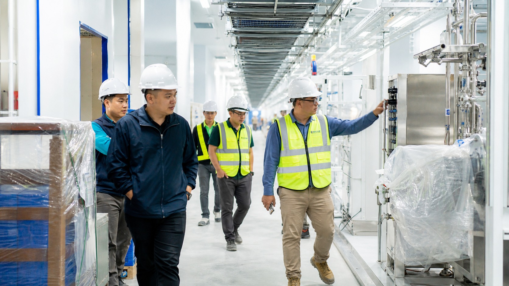 Technicians and contractors walking through a bright Malaysian high-tech factory construction corridor with cable trays, pallets, and industrial equipment